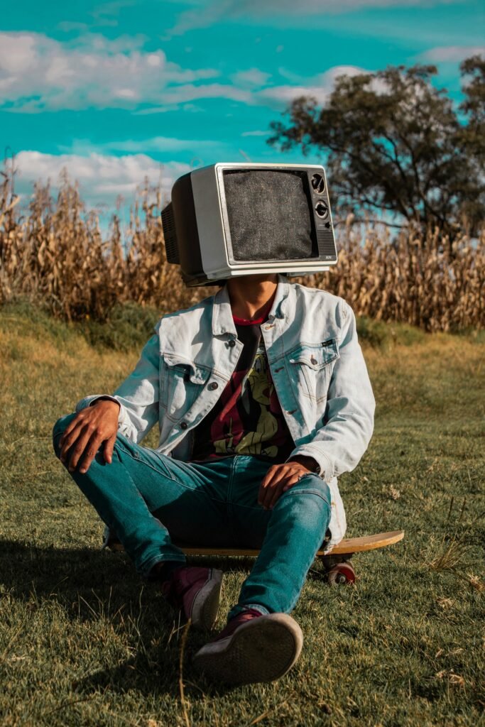 A person with a retro TV head sitting on a skateboard in a field.
