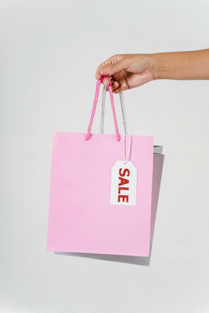 pexels-photo-5926247-5926247-1 Close-up of a hand holding a pink shopping bag with a sale tag against a white background.